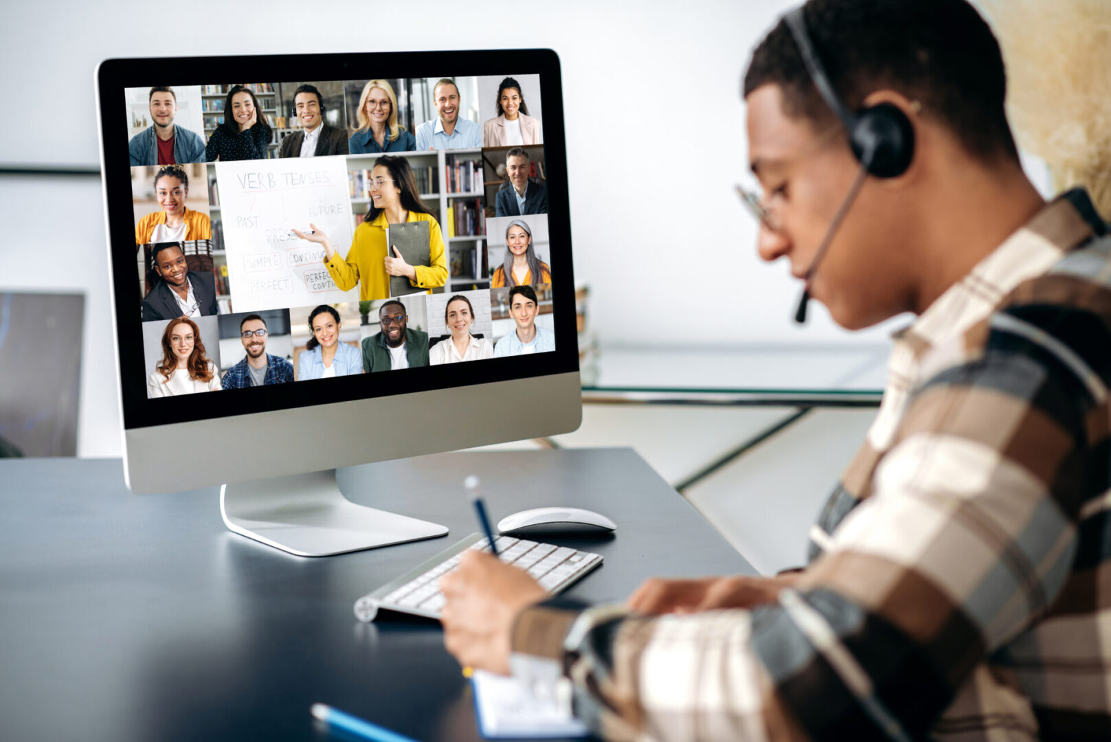 Successful mixed race clever male student learning distantly. At laptop screen female teacher and students. Teacher conducts online lecture, male student with headset takes notes. Video call